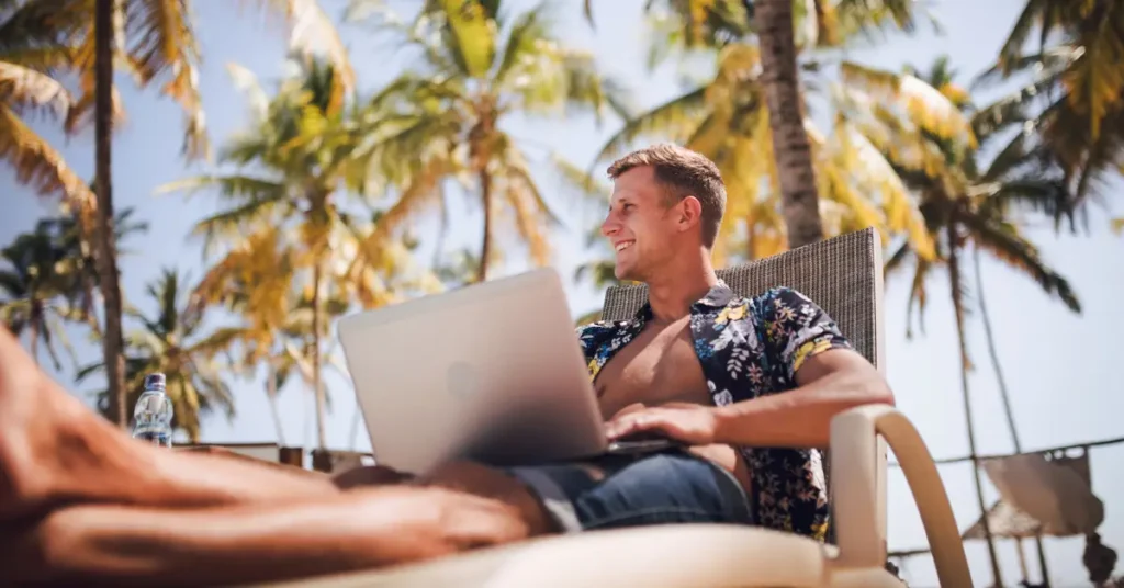 A man using a laptop while relaxing at a tropical beach resort, illustrating the freedom of the digital nomad lifestyle enabled by the Five Flag Theory.