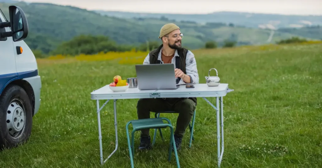 A digital nomad working on a laptop at an outdoor table in a scenic field, representing remote work freedom and the Five Flag Theory lifestyle.