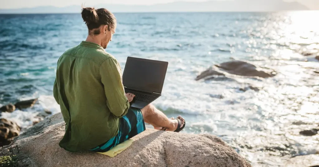 A person sitting on coastal rocks with a laptop, working beside the ocean to represent global mobility and location-independent living.